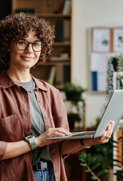 Young successful manager in casualwear standing in front of camera in coworking space and using laptop while analyzing online data